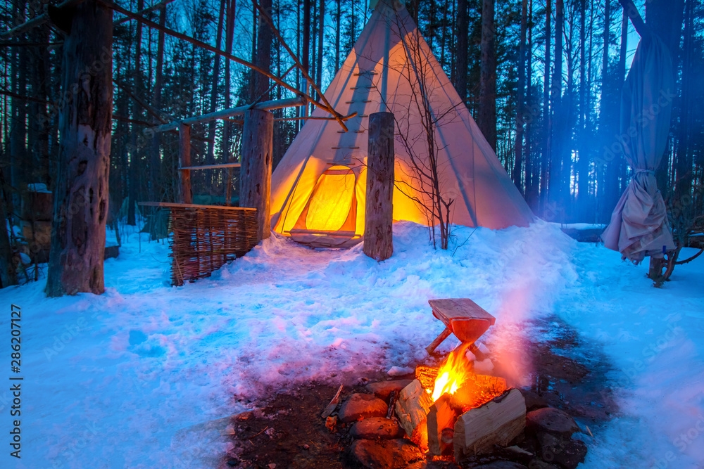 Tipi in winter forest. National Indian house. Nomadic lifestyle. The Lodge  is lit from the inside. Campfire in front of the tent. Eco-tourism. Photos  | Adobe Stock