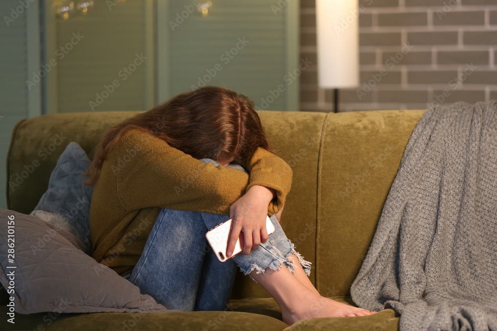 Offended teenage girl with mobile phone on sofa at night