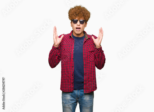 Young handsome man with afro hair wearing sunglasses celebrating mad and crazy for success with arms raised and closed eyes screaming excited. Winner concept photo