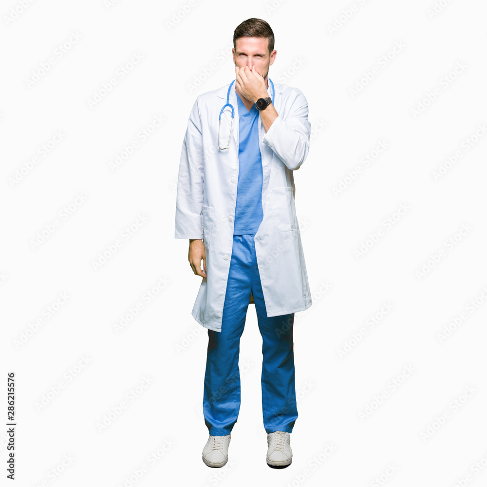 Handsome doctor man wearing medical uniform over isolated background smelling something stinky and disgusting, intolerable smell, holding breath with fingers on nose. Bad smells concept. Handsome doctor man wearing medical uniform over isolated background smelling something stinky and disgusting, intolerable smell, holding breath with fingers on nose. Bad smells concept.