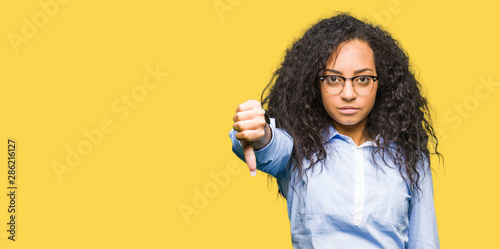 Young beautiful business girl with curly hair wearing glasses looking unhappy and angry showing rejection and negative with thumbs down gesture. Bad expression.