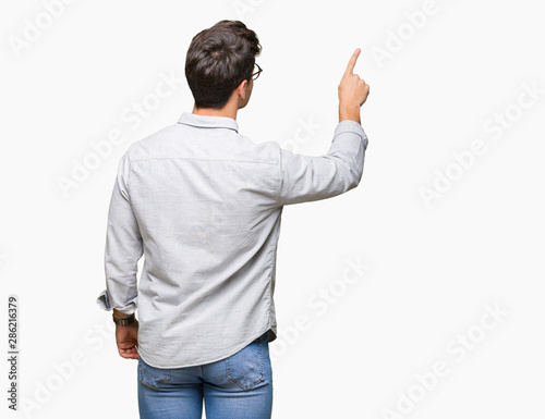 Young handsome man wearing glasses over isolated background Posing backwards pointing behind with finger hand