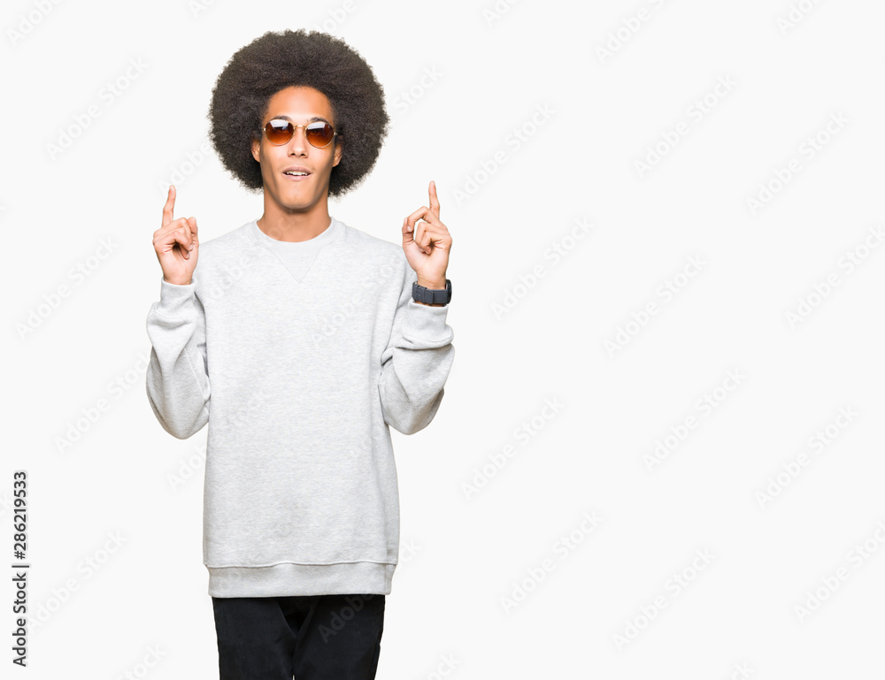 Young african american man with afro hair wearing sunglasses amazed and surprised looking up and pointing with fingers and raised arms. Young african american man with afro hair wearing sunglasses amazed and surprised looking up and pointing with fingers and raised arms.
