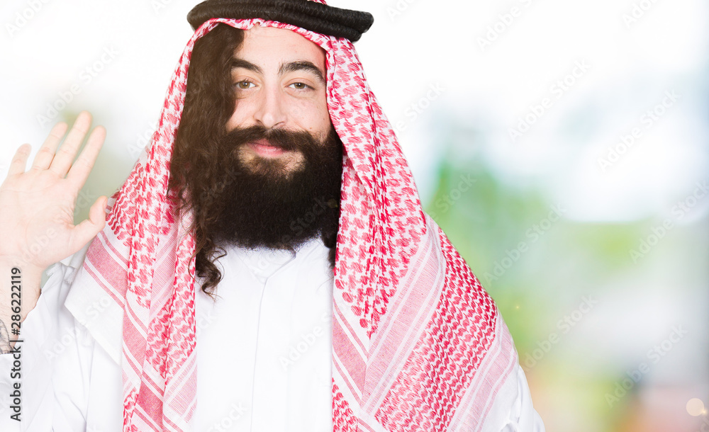 Arabian business man with long hair wearing traditional keffiyeh scarf Waiving saying hello happy and smiling, friendly welcome gesture