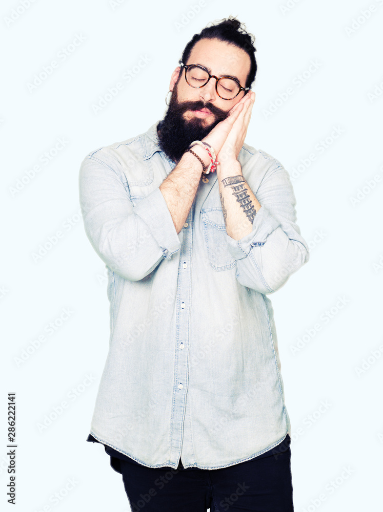 Young hipster man with long hair and beard wearing glasses sleeping tired dreaming and posing with hands together while smiling with closed eyes. Young hipster man with long hair and beard wearing glasses sleeping tired dreaming and posing with hands together while smiling with closed eyes.