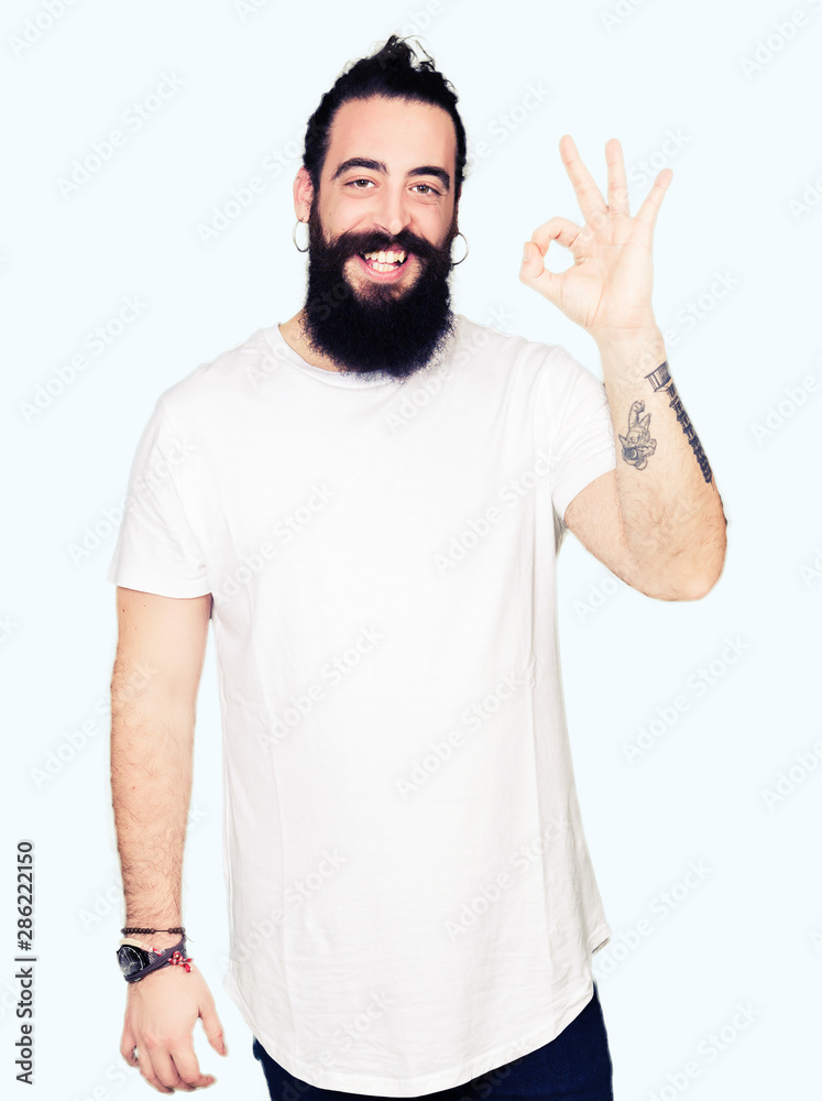 Young hipster man with long hair and beard wearing casual white t-shirt smiling positive doing ok sign with hand and fingers. Successful expression. Young hipster man with long hair and beard wearing casual white t-shirt smiling positive doing ok sign with hand and fingers. Successful expression.
