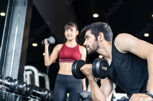 Young Caucasian handsome and pretty Asian girl are lifting the weights in gym or fitness club. They both have very strong and good body shape. Seen side of the man is looking straight in the mirror. © Kawee