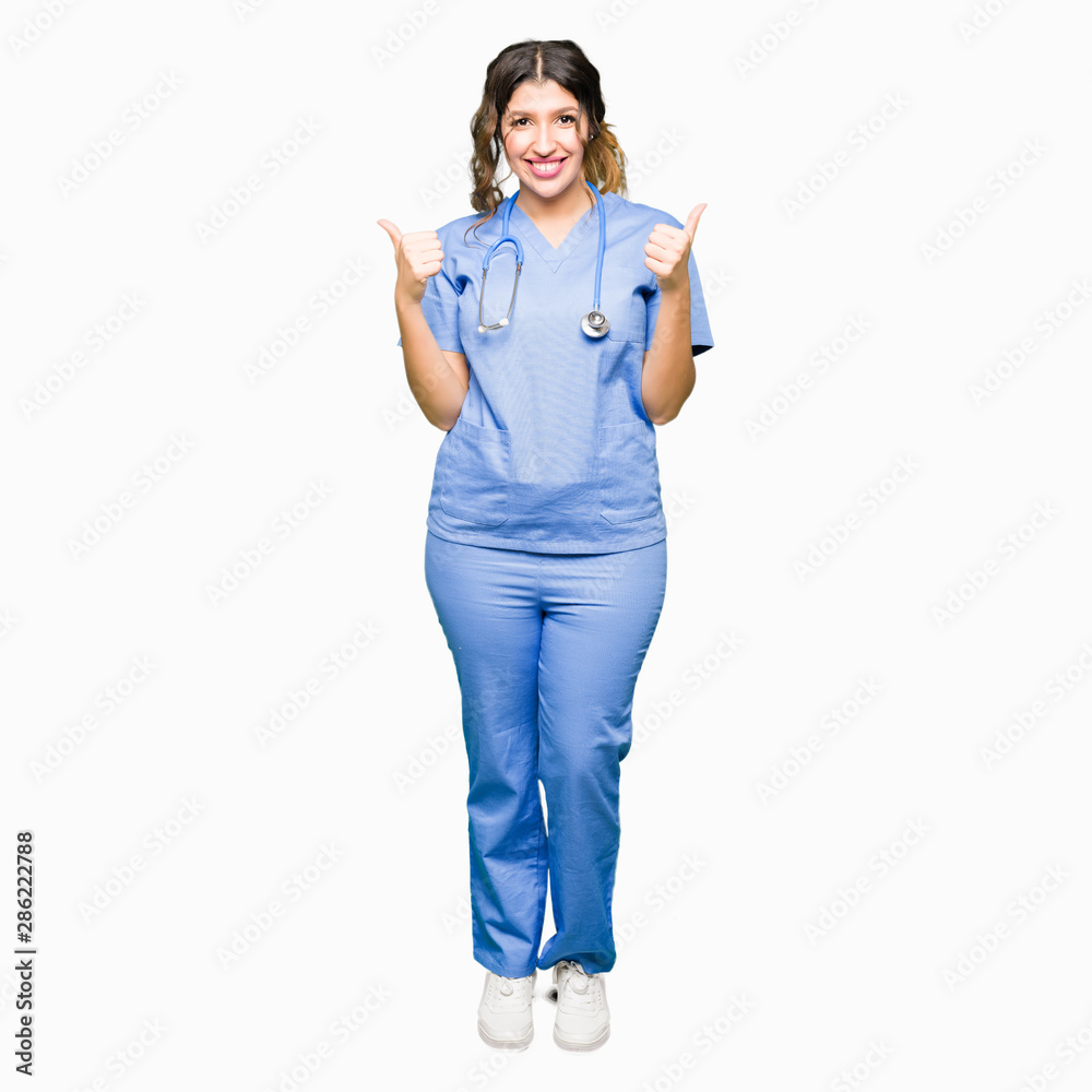 Young adult doctor woman wearing medical uniform success sign doing positive gesture with hand, thumbs up smiling and happy. Looking at the camera with cheerful expression, winner gesture. Young adult doctor woman wearing medical uniform success sign doing positive gesture with hand, thumbs up smiling and happy. Looking at the camera with cheerful expression, winner gesture.