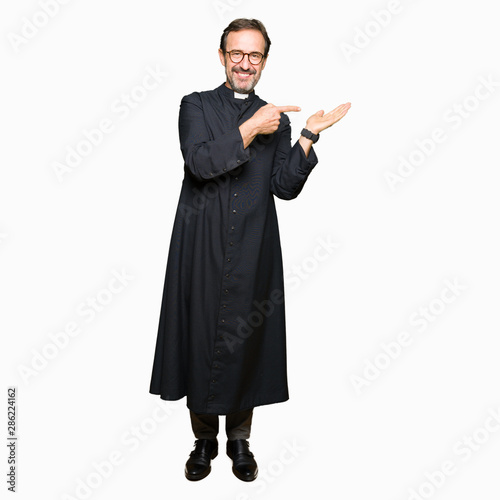 Middle age priest man wearing catholic robe amazed and smiling to the camera while presenting with hand and pointing with finger.