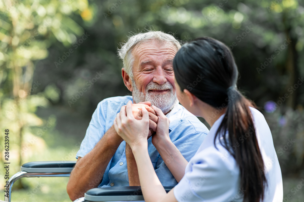 Naklejka premium Caring nurse with senior man sitting on wheelchair in gaden. Asian woman, caucasian man. Asking questions.