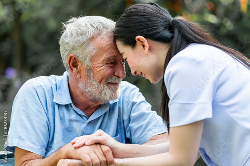 Caring nurse with senior man sitting on wheelchair in gaden. Asian woman, caucasian man. Heads against each other. Close up.