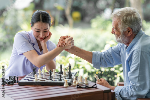Senior man playing chess with young nurse. White male with asian woman. Hand shake.