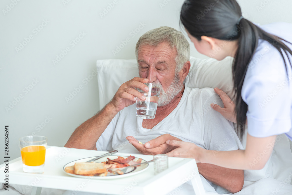 Senior man in bed having medication with nurse. Caucasian man with asian woman.  Drinking water after a pill.