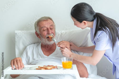 Senior man in bed having breakfast with nurse. Caucasian man with asian woman.  Feeding him beacon.