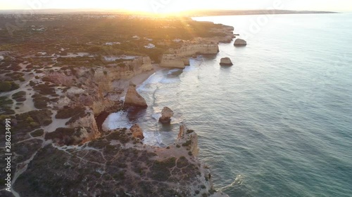 Aerial, reverse, drone shot, away from the Marinha beach, overlooking arches and rock formations, on the coast of Algarve, at sunrise, on a sunny, morning dawn, in Carvoeiro, Portugal