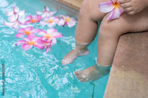 Fototapeta Naklejka Na Ścianę i Meble -  Baby feet in a swimming pool. Tropical flowers Frangipani Plumeria, Leelawadee floating in the water. Spa pool. Peace and tranquility. Spa concept.