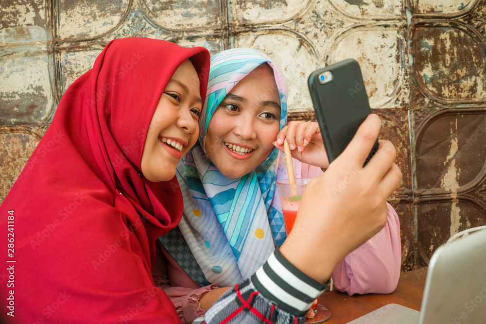 Fototapeta premium Happy and pretty Asian Indonesian girls in traditional Islamic hijab head scarf taking selfie together having fun at cafe in friendship and social media concept