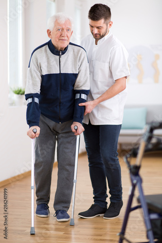 Elderly man walking on crutches and a helpful male nurse supporting him