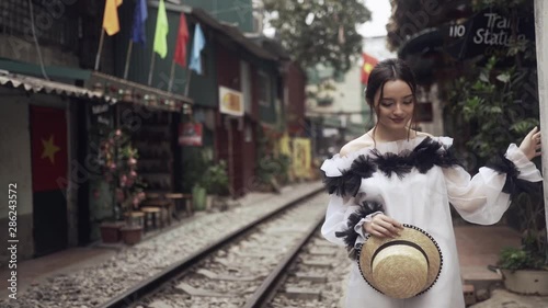 Beautiful and young fashion mestizo girl walking along the asian street with railway between old buildings and holding straw hat in hands. Journey concept in Vietnamese tourist destination in Hanoi. 