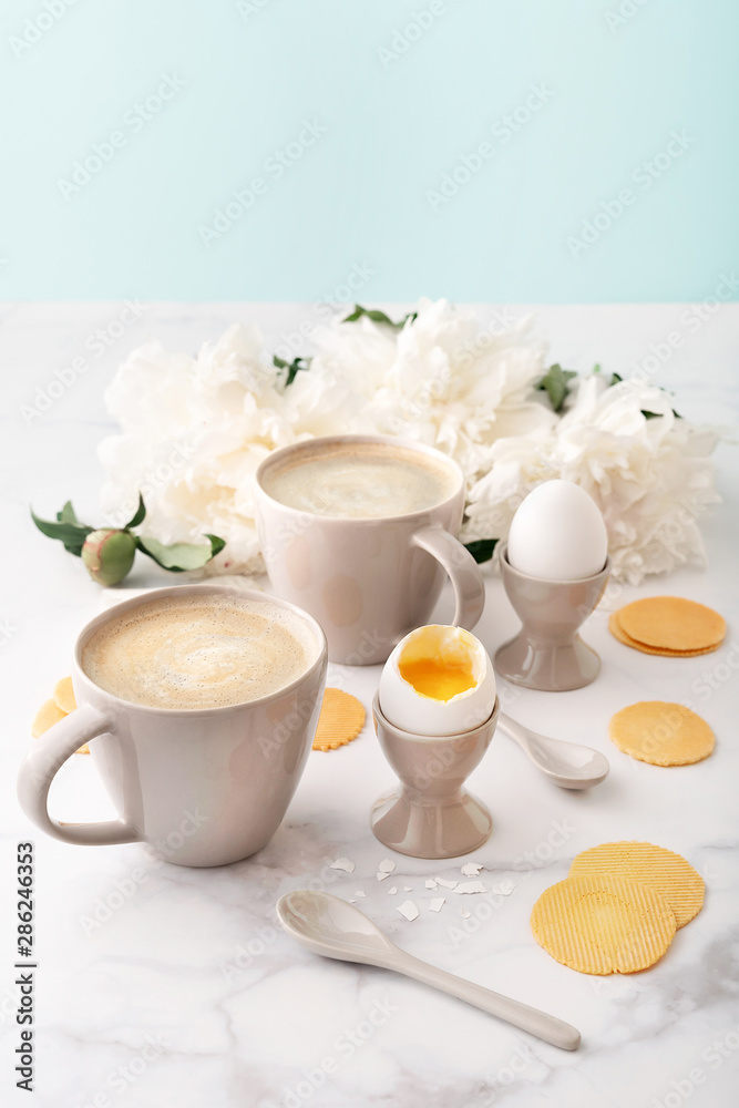 Soft-boiled eggs with liquide orange yolk in ceramic egg cups, two cups of coffee with ceramic spoons and thin crispy corn chips on background of beautiful white peony. Breakfast concept. Copy space.