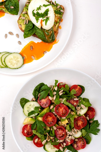 Sandwiches with poached eggs and salad with avocado, cucumbers, cherry tomatoes, spinach leaves, parsley and seeds. Breakfast food on a white background. Top view.