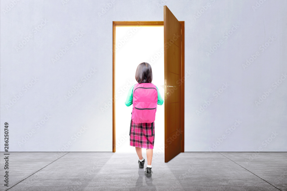 Fototapeta premium Rear view of Asian little girl with a backpack walking through the door
