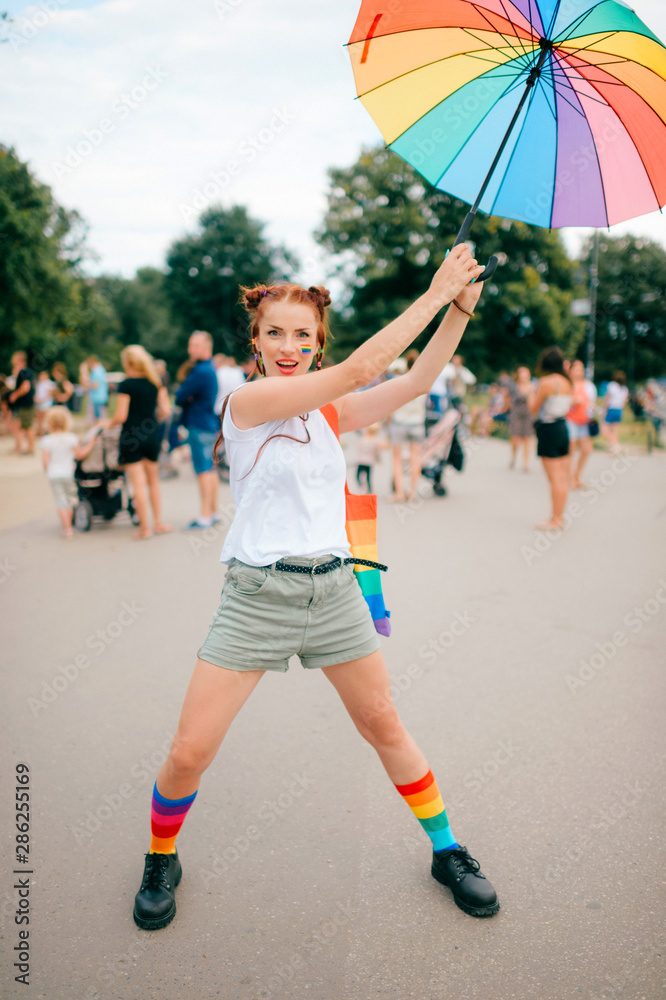 Obraz premium bisexual girl with lgbt gay pride flag on her face, colorful umbrella, bag and mismatched socks posing on street with people on background.