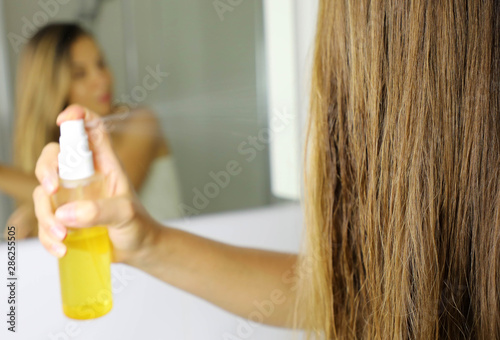 Woman applying oil mask spray on hair in front of a mirror. Haircare concept. Focus on hair.