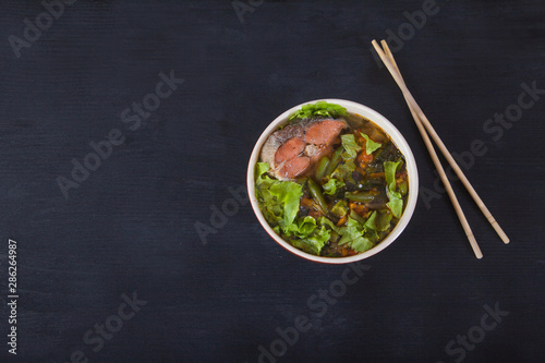 Traditional japanese soup on a red napkin on a black background. A female hand takes out tofu cheese with chopsticks. Top view. Horizontally.