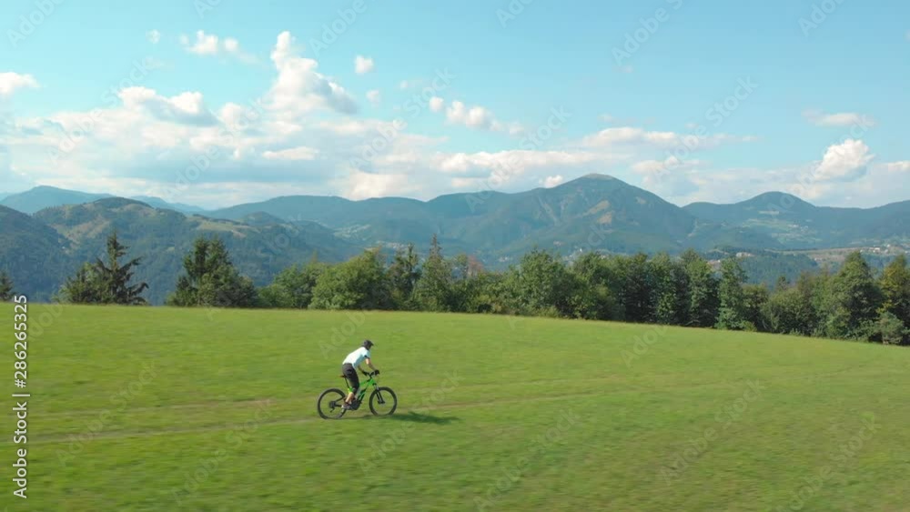 AERIAL: Unrecognizable man speeds down a single trail crossing a large meadow on a green electric bicycle. Athletic male tourist enjoying his day off by downhill cycling in the scenic countryside. AERIAL: Unrecognizable man speeds down a single trail crossing a large meadow on a green electric bicycle. Athletic male tourist enjoying his day off by downhill cycling in the scenic countryside.