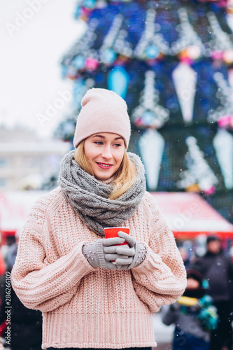 cute young girl wearing hat and pink sweater. christmas market lights bokeh background. Woman drinking hot chocolate in winter morning in christmas market.