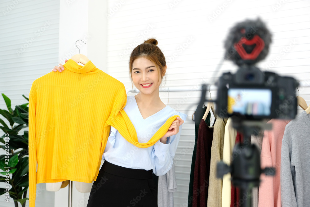Youn asian woman fashion blogger showing cloth with smiling face while recording new content for vlog and fashion online shopping with camera at home studio