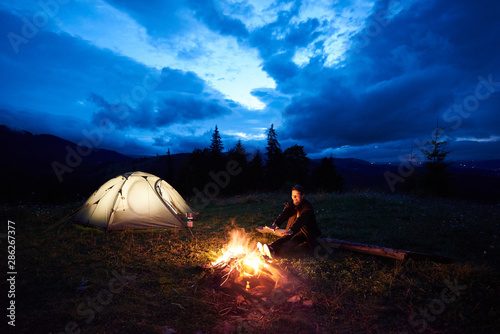 Young woman traveller enjoying at night camping in the mountains  sitting near burning campfire and illuminated tourist tent under beautiful evening cloudy sky. Tourism  outdoor activity concept