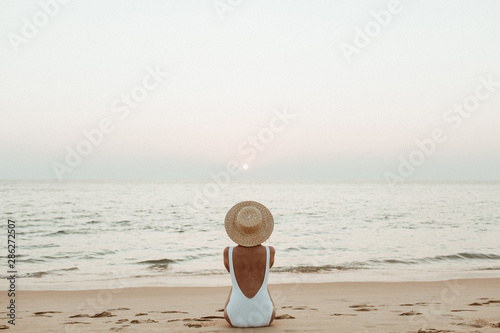 Summer vacation fashion concept. Young, tanned woman wearing a beautiful white swimsuit with a straw hat is sitting and relaxing on tropical beach with white sand and is watching sunset and sea. photo