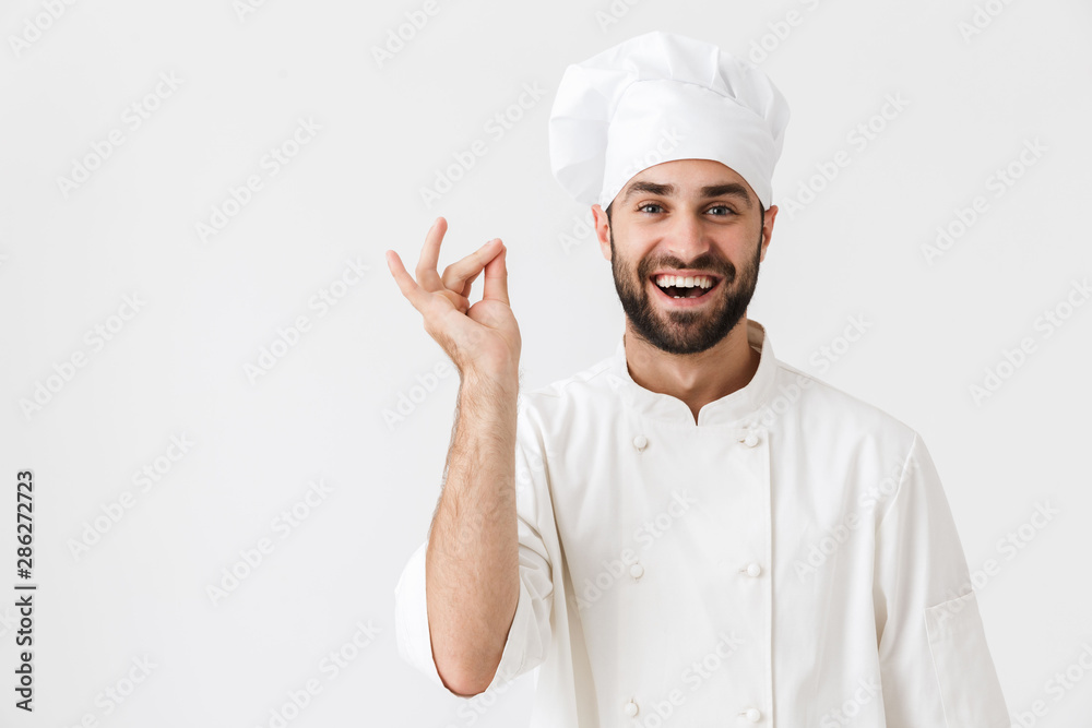 Cheerful pleased young chef posing isolated over white wall background in uniform. Cheerful pleased young chef posing isolated over white wall background in uniform.