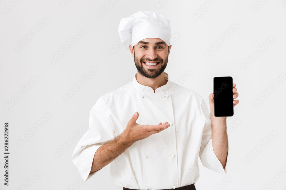 Positive smiling young chef posing isolated over white wall background in uniform holding mobile phone showing empty display. Positive smiling young chef posing isolated over white wall background in uniform holding mobile phone showing empty display.
