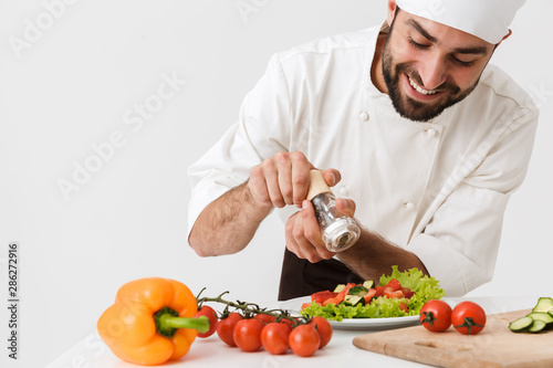 Pleased positive happy young chef isolated over white wall background in uniform cooking with fresh vegetables.