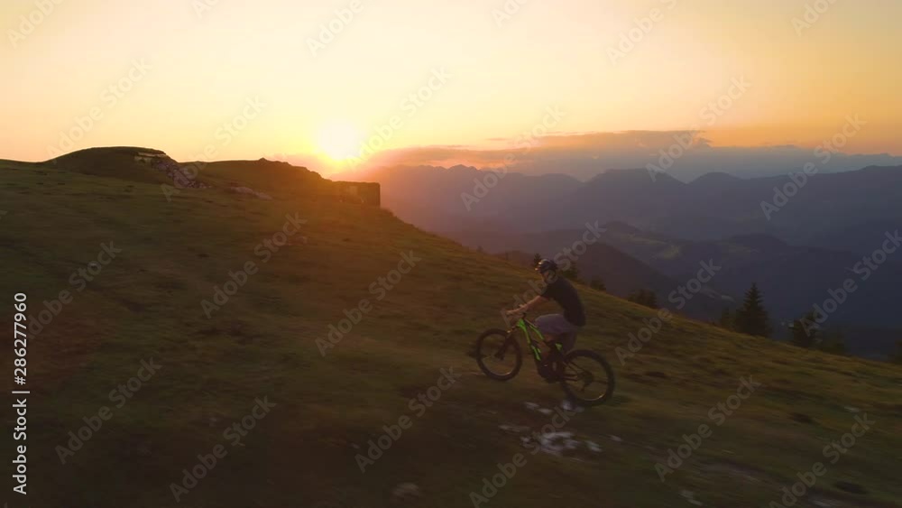 DRONE: Golden sunset illuminates the way for a young mountain biker pedalling uphill.Flying along an athletic male tourist riding his electric bike up a steep grassy hill early in the morning. DRONE: Golden sunset illuminates the way for a young mountain biker pedalling uphill.Flying along an athletic male tourist riding his electric bike up a steep grassy hill early in the morning.