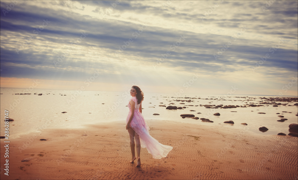 Young slim beautiful smiling brunette woman in pink dress posing on sunset ocean beach. Young slim beautiful smiling brunette woman in pink dress posing on sunset ocean beach.