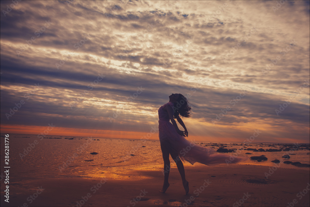 Silhouette of young slim beautiful smiling brunette woman in a long pink dress posing on sunset ocean beach. 