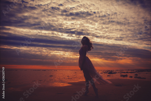 Silhouette of young slim beautiful smiling brunette woman in a long pink dress posing on sunset ocean beach. 