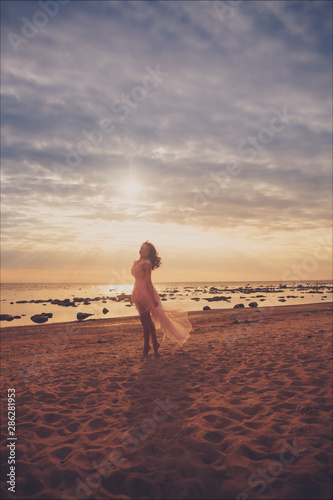 Young slim beautiful smiling brunette woman in pink dress posing on sunset ocean beach.  © Алина Троева