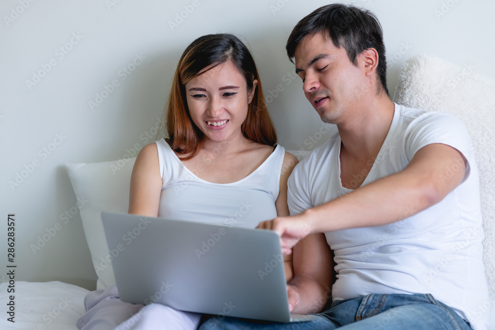 Pregnant woman in bed with man. Asian woman with white man. Using laptop. Mix race relationship concept. Happy smile.