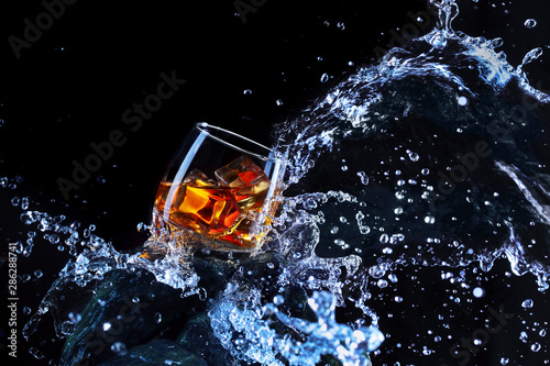 Glass of whiskey with ice .Water splash the glass.Creative photo glass of whiskey on stone with fog and black background.Copy space.Advertising shot