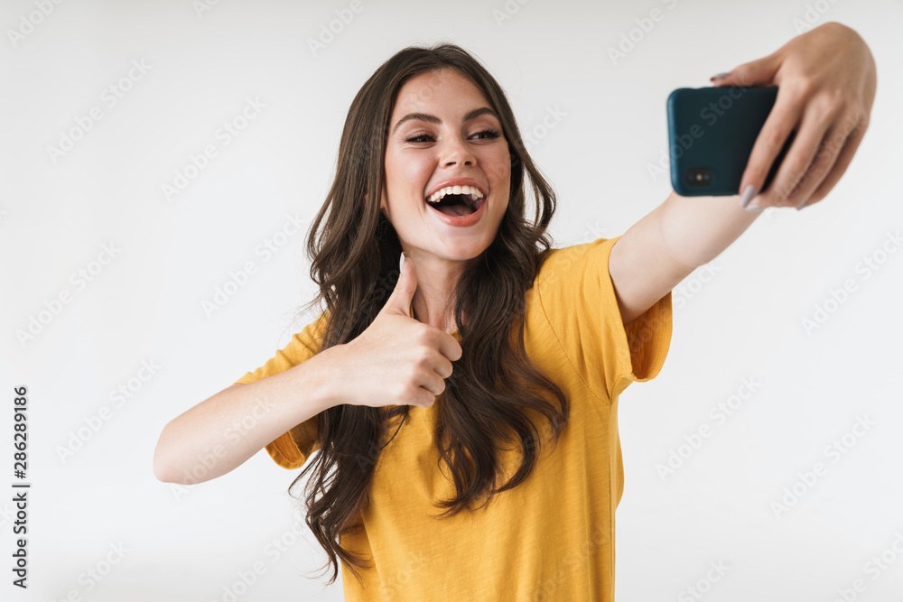 Image of caucasian brunette woman laughing and showing thumb up while taking selfie photo on cellphone Image of caucasian brunette woman laughing and showing thumb up while taking selfie photo on cellphone