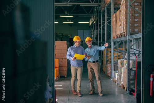CEO going around warehouse with supervisor and talking analyzing sale statistics. Younger man holding folder with data while older one holding tablet and pointing at boxes. Both having yellow helmets.