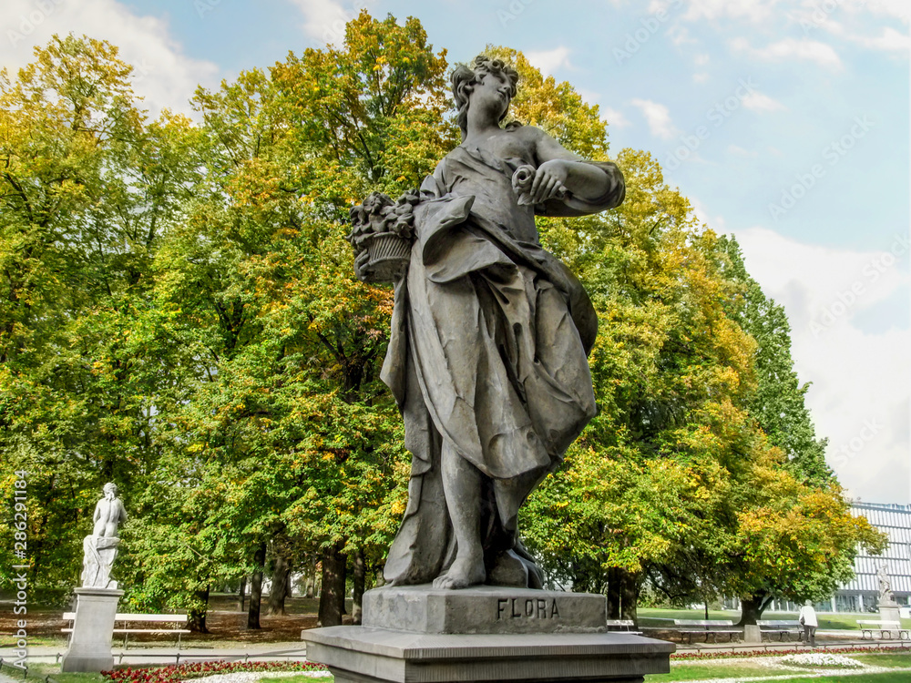 Sandstone sculpture of the goddess Flora in the Rococo style in Saxon Garden in Warsaw, Poland. Park sculpture of the ancient Roman goddess on a background of yellow-green foliage of trees, close-up