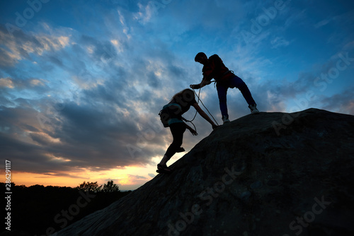 Two climbers getting top of cliff, one holding hand of his partner assisting to make last step to top. Teamwork couple helping hand trust help, silhouettes in mountains. Amazing heaven on background.