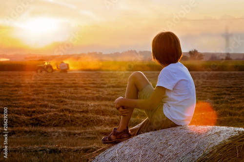 Happy child, watching tractor working in field on sunset, sitting on haystack