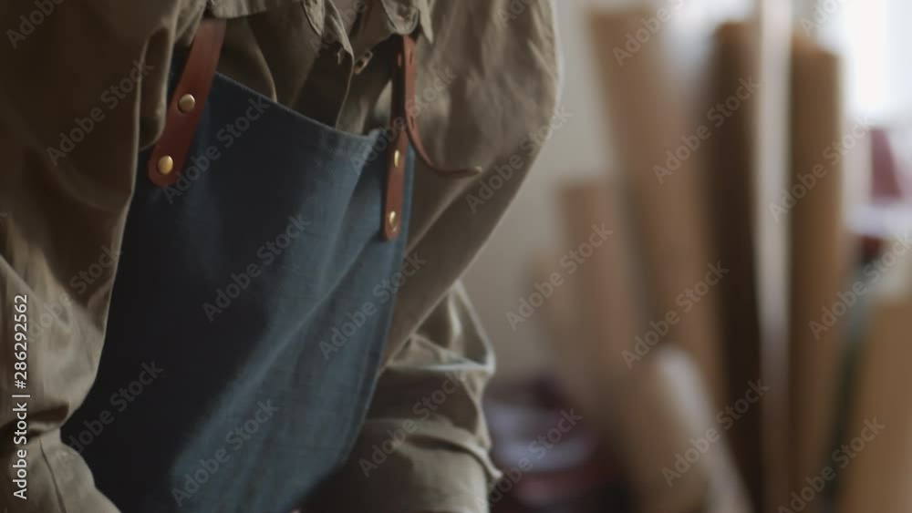Tilt down of Caucasian craftswoman in apron standing at worktop and smoothing piece of brown leather with her hands